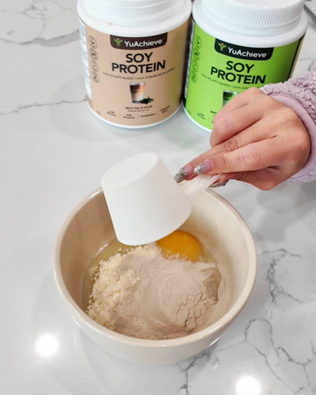 A top-down view of a person's hand with silver glitter nails pouring a white scoop of YuAchieve soy protein powder into a ceramic mixing bowl containing a cracked egg and dry ingredients. In the background, two large tubs of YuAchieve Soy Protein in Milk Tea and Matcha Latte flavors are visible on a white marble countertop, capturing the preparation stage of a high-protein pancake recipe.