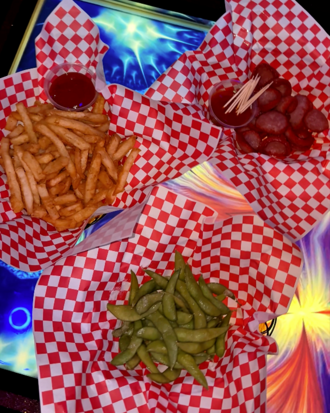 Baskets of crispy french fries with ketchup, savory sliced sausages, and seasoned edamame served in red and white checkered paper on a glowing interactive lounge table at ET KTV Markham