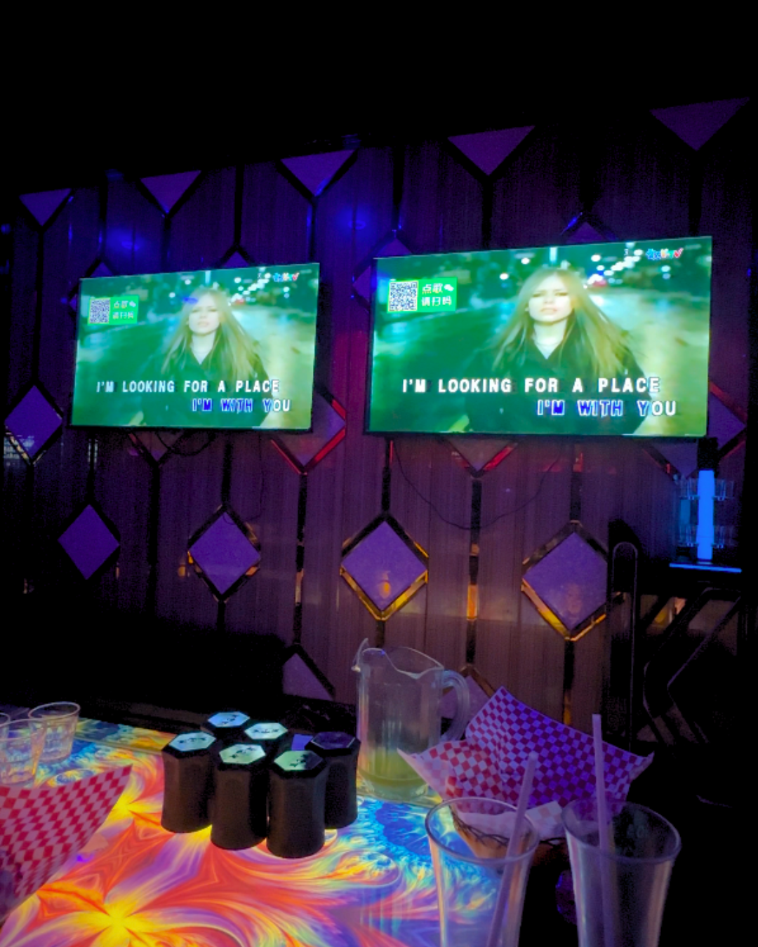 Dual flat screen TVs playing an Avril Lavigne music video with karaoke lyrics over a colorful glowing display table at Entertainment Tonight KTV Markham.