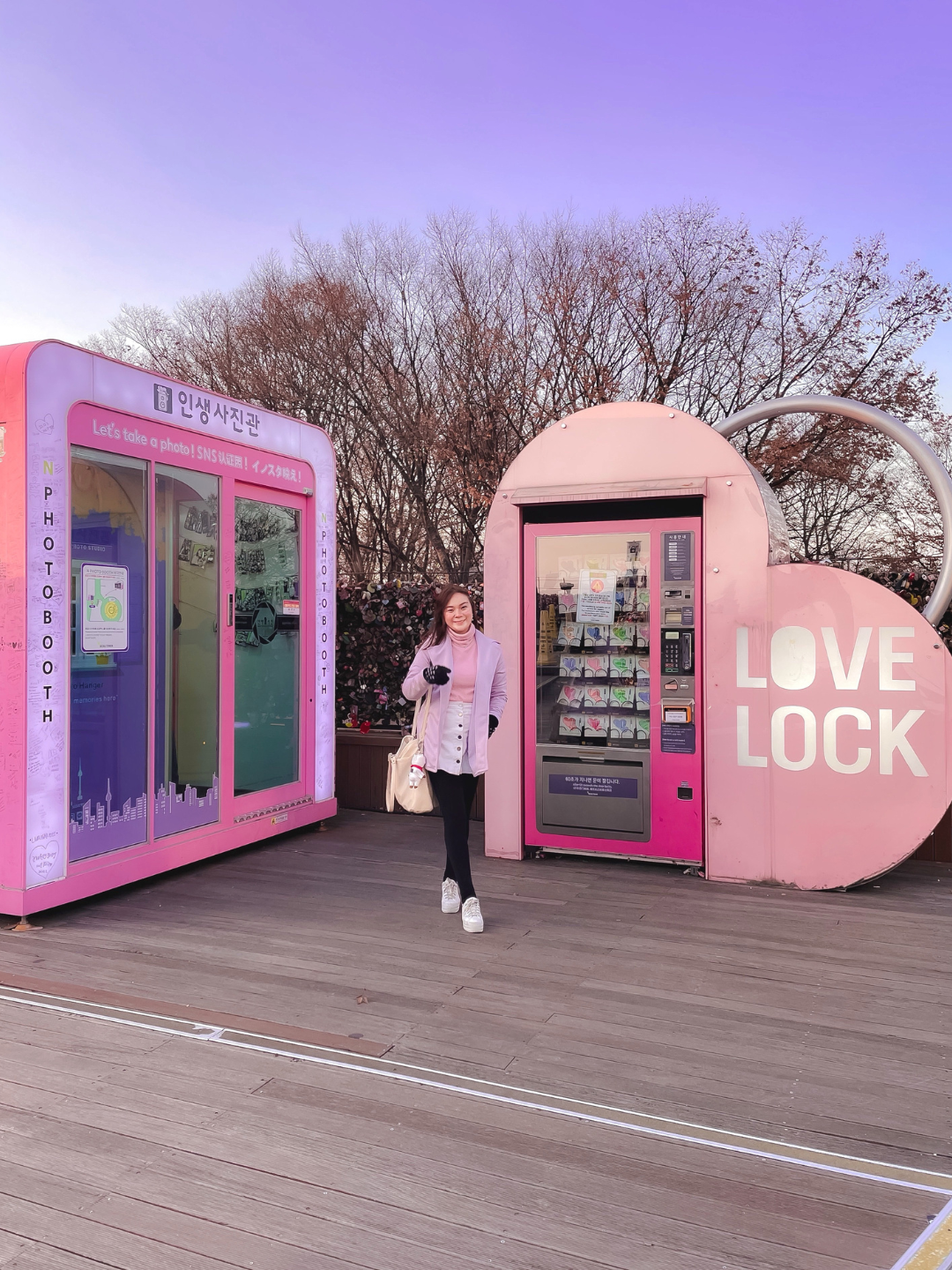 A traveler posing on the wooden observation deck at N Seoul Tower between a bright pink photo booth and a heart-shaped love lock vending machine. A practical location spot guide for ARMYs looking for aesthetic photo zones on Namsan Mountain.