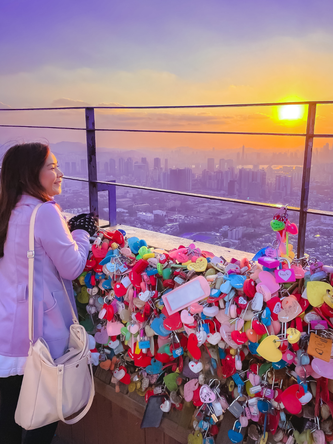 Roxanne Lacap - A Toronto based lifestyle content creator in a pastel lilac coat looking out at a stunning golden sunset over the Seoul city skyline, standing by the iconic colorful love locks railing at N Seoul Tower. An aesthetic travel inspiration shot featured in a BTS Festa Seoul itinerary.
