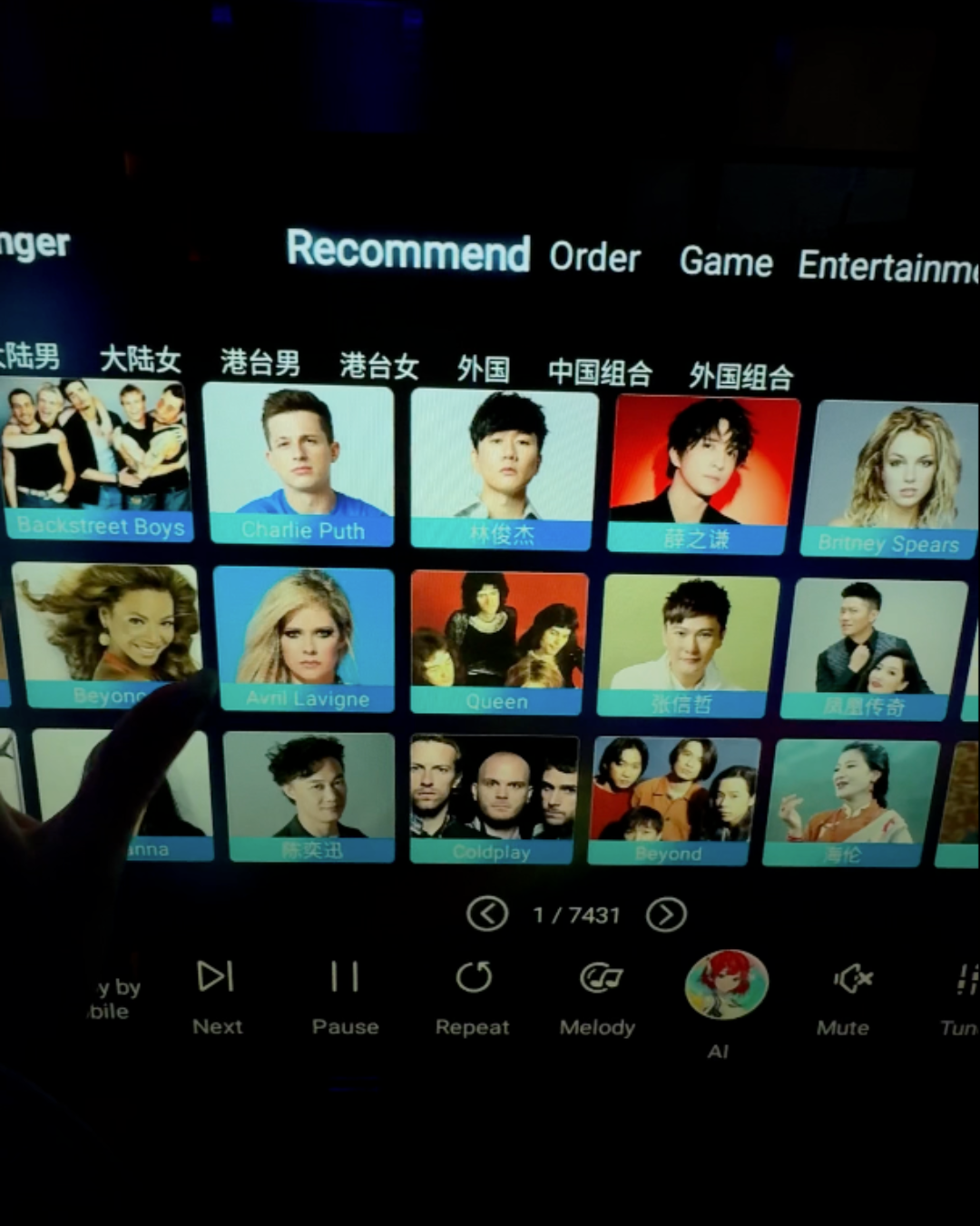 A hand selecting an artist on a large digital touchscreen karaoke song selection monitor featuring pop and international musicians at ET KTV Markham