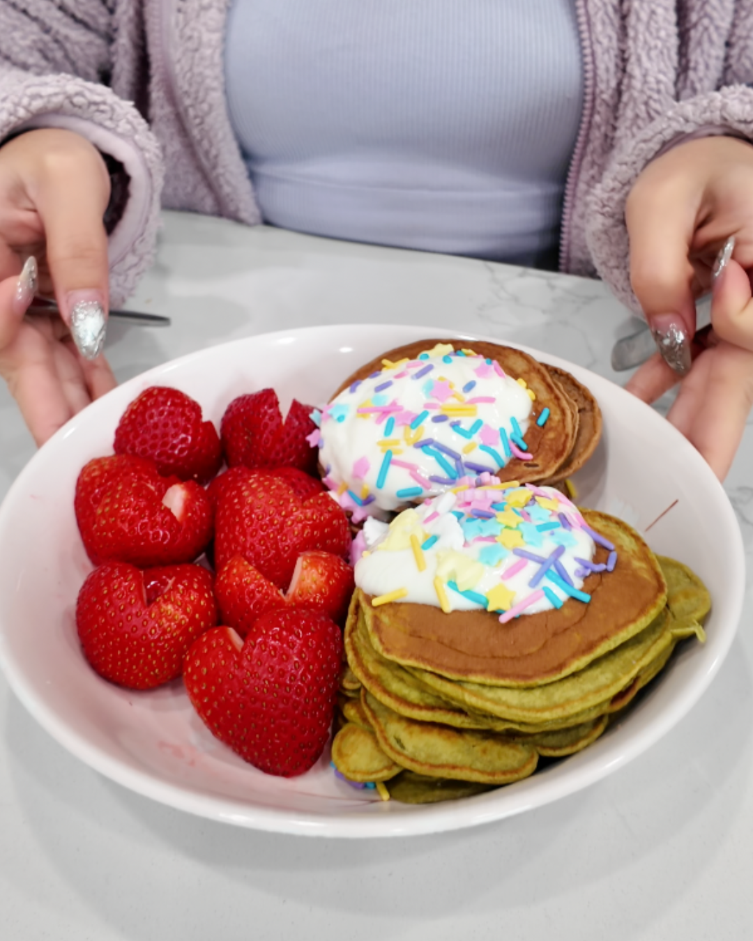 A close-up, top-down view of a person wearing a purple fuzzy jacket holding a large white bowl of whimsy protein pancakes. The bowl features a stack of golden-brown matcha-infused pancakes topped with a generous dollop of white yogurt and a vibrant mix of pastel blue, pink, and yellow star-shaped sprinkles. Beside the pancakes are several fresh, bright red strawberries meticulously carved into heart shapes, all presented on a clean white marble countertop