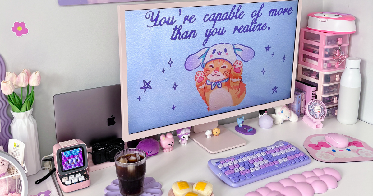 A wide-angle view of a cozy pink and purple aesthetic desk setup featuring an LG monitor with a cat wallpaper, a matching lavender cat keyboard, a pink cloud wrist rest, a Divoom speaker, and a glass of iced coffee.