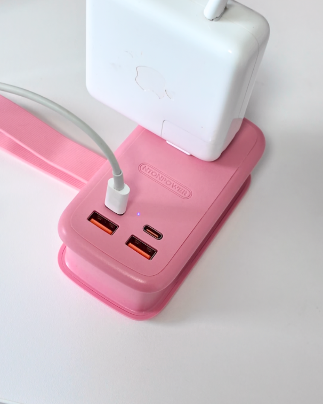 A high angle shot of a large white Apple MacBook charger and a thin grey USB cable plugged directly into the pink NTONPOWER desktop charging hub on a white surface.