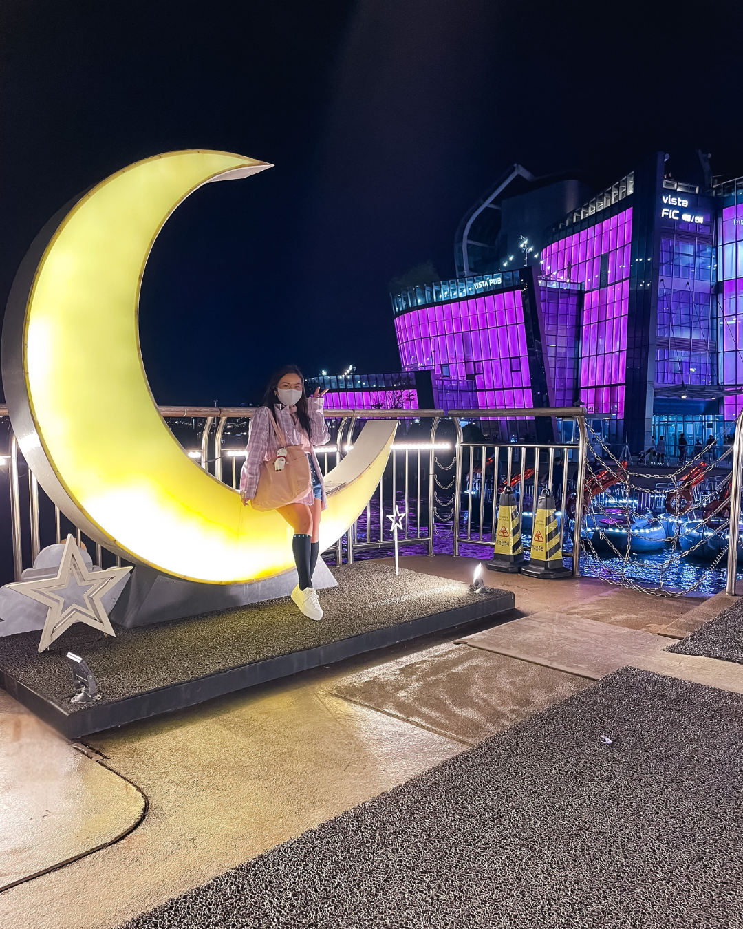 Roxanne Lacap sitting on a glowing crescent moon installation at the Han River in Seoul Korea at night with purple lights in the background