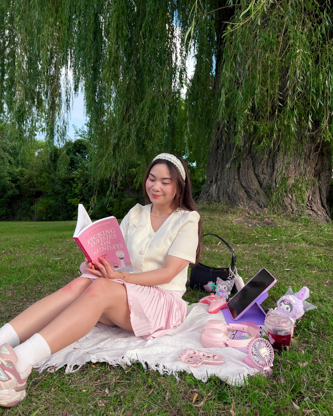 Roxanne Lacap Toronto lifestyle creator reading a pink romcom book outside on a blanket under a willow tree with cute kawaii accessories whimsy spring activities