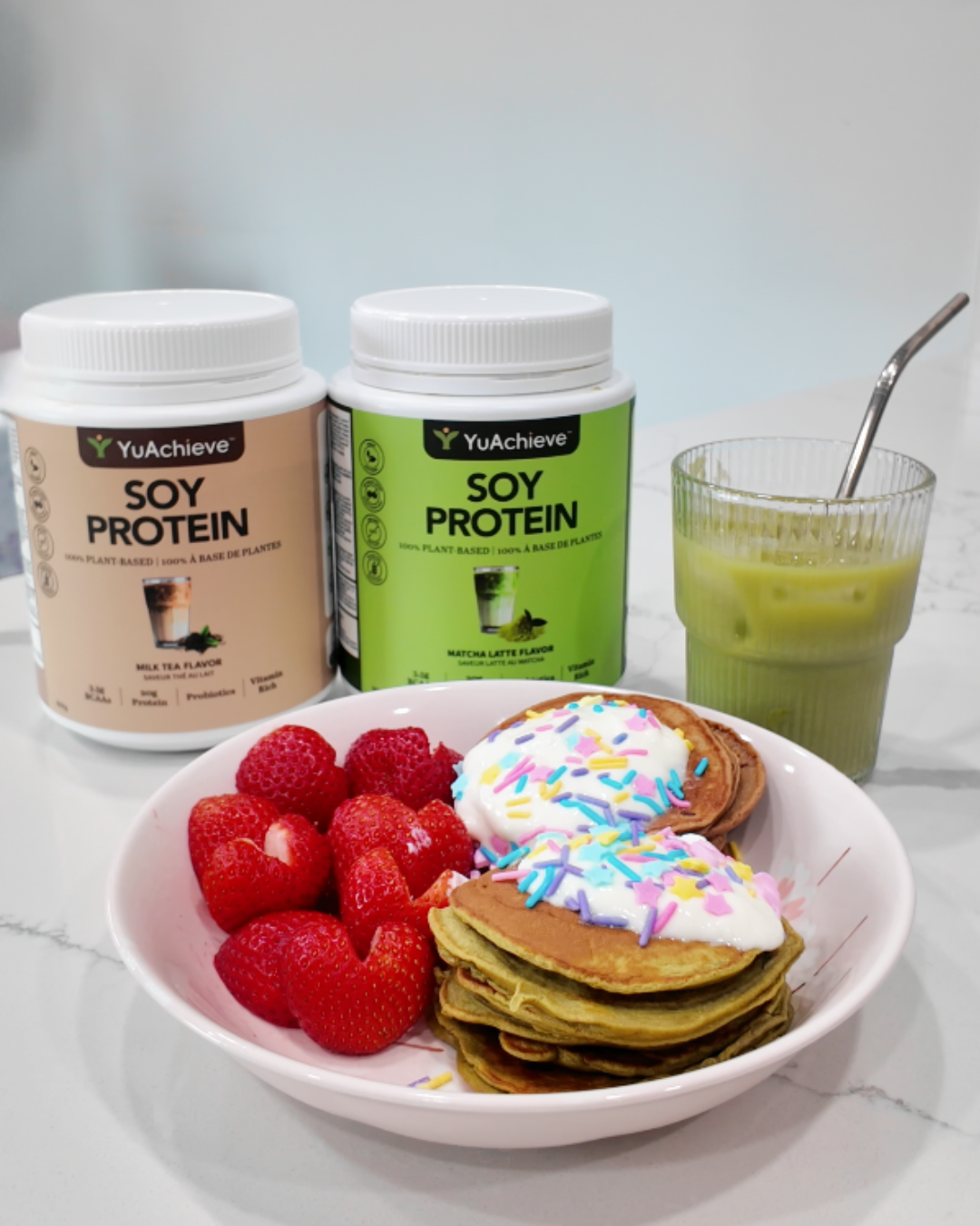 A stack of green matcha protein pancakes in a pink bowl topped with white yogurt and pastel star sprinkles, served alongside heart-shaped halved strawberries. In the background, two tubs of YuAchieve Soy Protein in Milk Tea and Matcha Latte flavors sit next to a ribbed glass of iced matcha latte with a metal straw on a white marble countertop