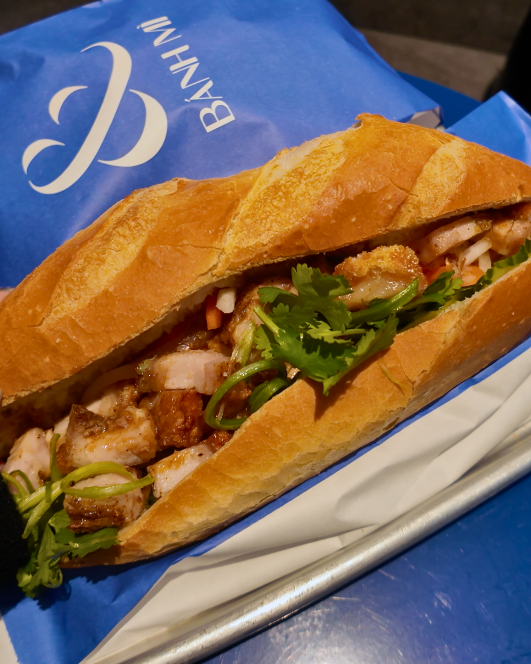 Close up view of the & crackle sandwich at AndBanhMi Toronto showcasing a perfectly crispy baguette loaded with roasted pork belly, fresh cilantro, and pickled carrots next to a blue branded paper bag