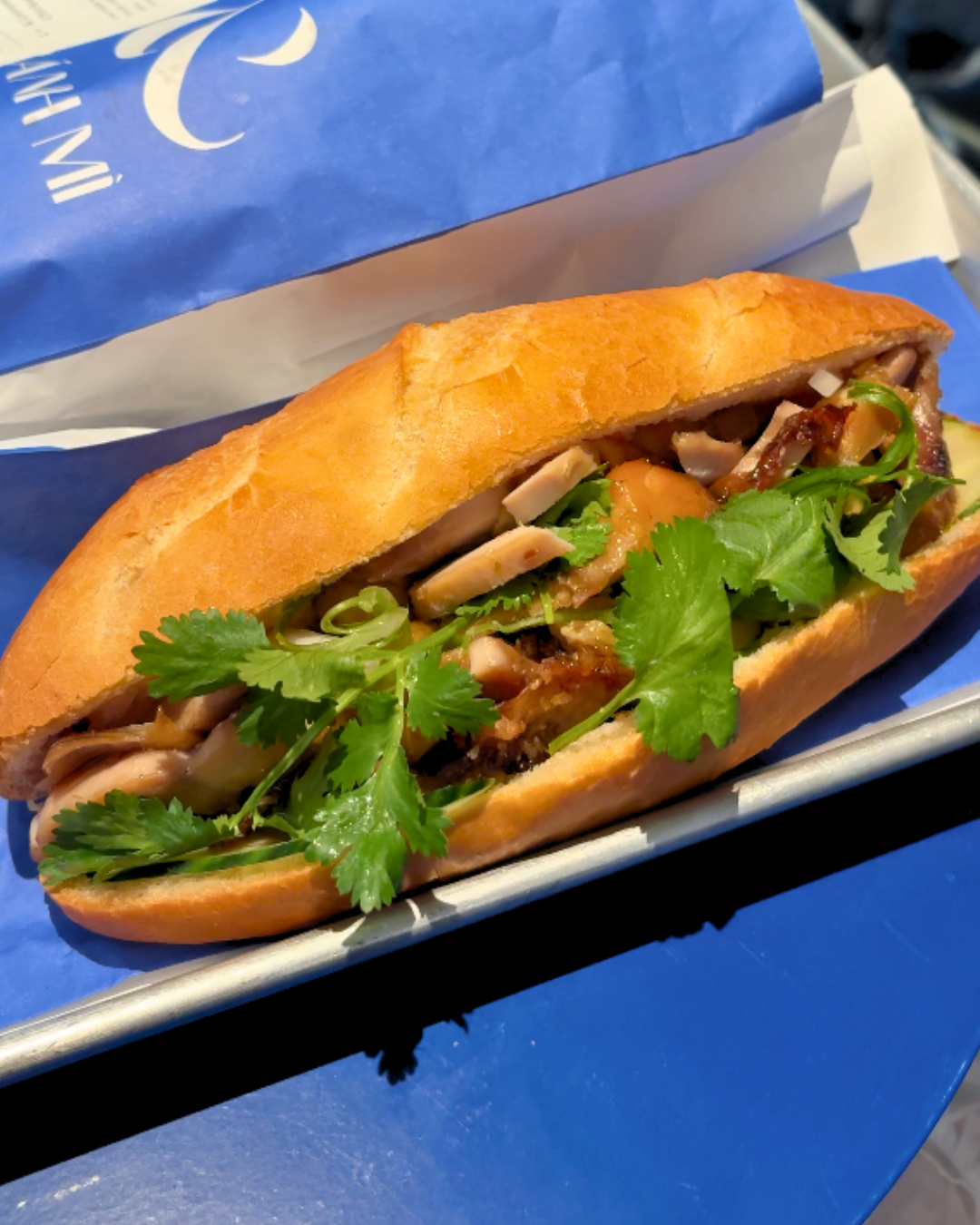 Close up view of the & lemongrass chicken sandwich at AndBanhMi Toronto featuring crispy fresh bread filled with savory chicken, cilantro, and pickled vegetables on a metal tray