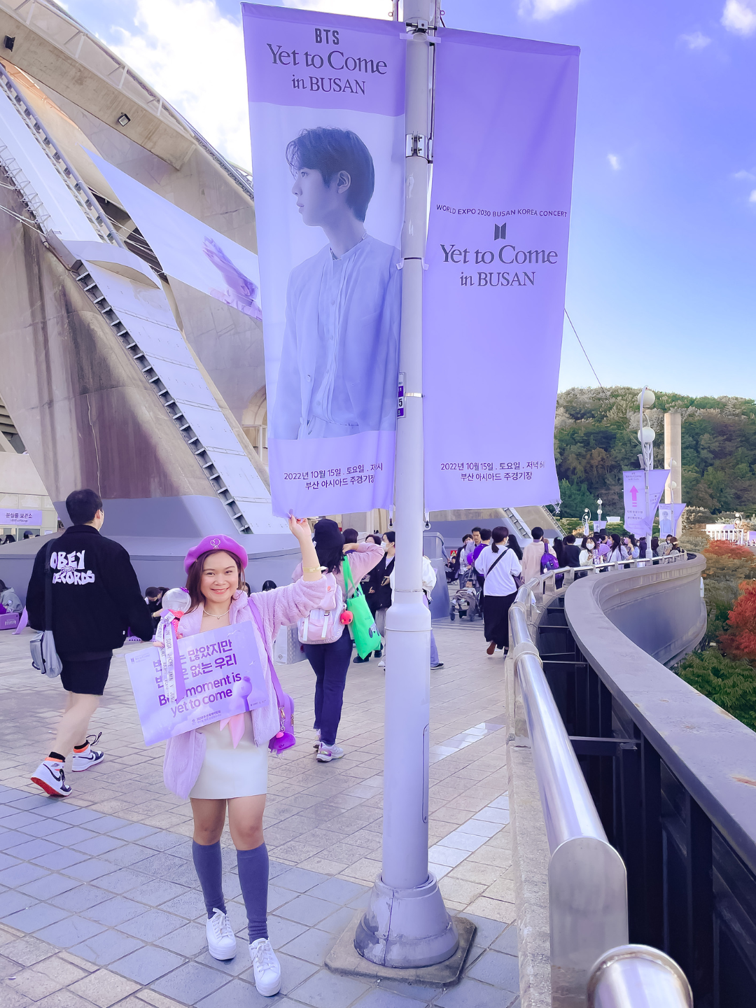 BTS ARMY in a purple beret and pink jacket holding up an official concert slogan banner outside the Busan Asiad Main Stadium, posing next to a tall light pole decorated with large purple BTS concert banners amid crowds of fans. An essential visual for an ARMY travel guide to the ARIRANG tour and Festa in Busan