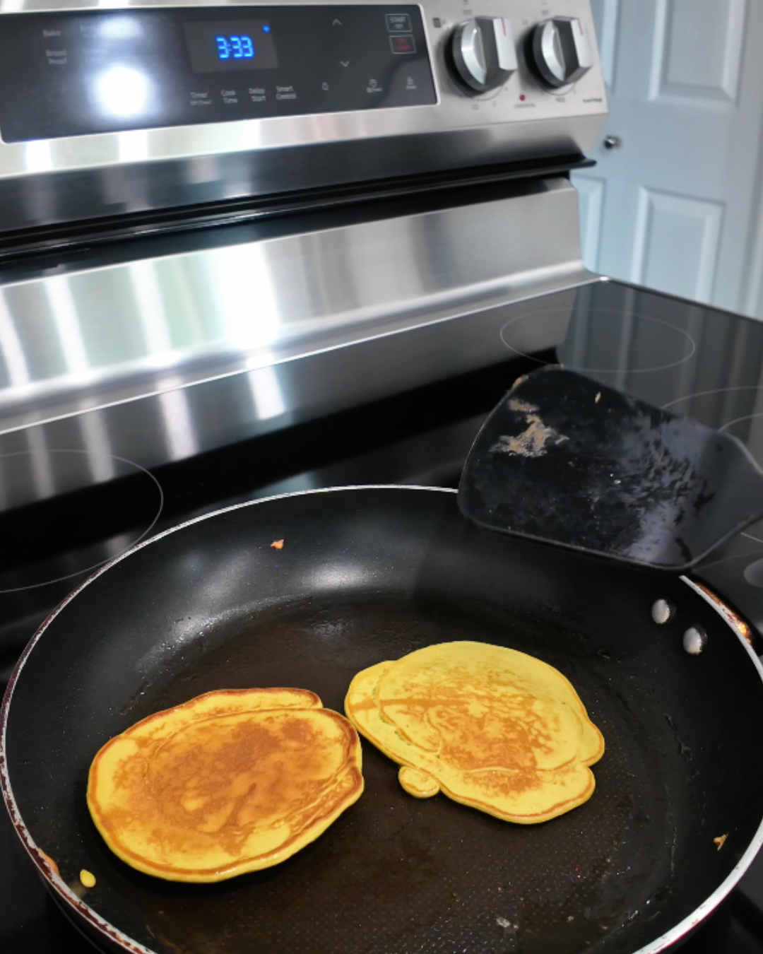 A close-up shot of several small, round matcha protein pancakes cooking on a black non-stick griddle. A person's hand with silver glitter nails is using a stainless steel spatula to flip a golden-brown pancake, showing the vibrant green matcha interior. The stovetop setting captures the cooking process of a healthy, high-protein breakfast.