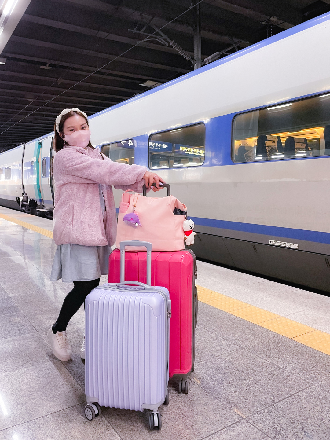 BTS ARMY A lifestyle content creator in a pastel pink plush jacket and matching face mask standing on a train platform next to a sleek white and blue KTX bullet train, holding a bright pink suitcase and a lilac carry-on bag with a purple whale plush keychain. Perfect for a travel guide detailing the road trip from Seoul to Busan for the BTS ARIRANG tour.