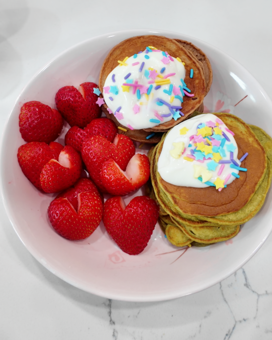 A beautiful flat-lay of a finished high-protein breakfast featuring a stack of matcha protein pancakes topped with creamy white yogurt and colorful pastel star sprinkles. A pink bowl on a marble countertop holds fresh strawberries sliced into heart shapes. In the background, two containers of YuAchieve Soy Protein and an iced matcha latte in a ribbed glass complete the aesthetic morning meal