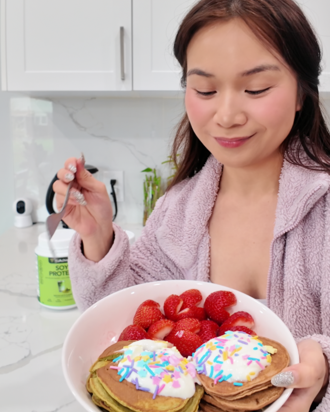 A vertical flat-lay of a completed protein pancake meal on a white marble surface. A pink bowl contains a stack of matcha pancakes topped with white yogurt and colorful pastel sprinkles, paired with fresh strawberries cut into heart shapes. To the left, two tubs of YuAchieve Soy Protein in Milk Tea and Matcha flavors are displayed, and to the right, a tall ribbed glass holds an iced matcha latte with a metal straw.