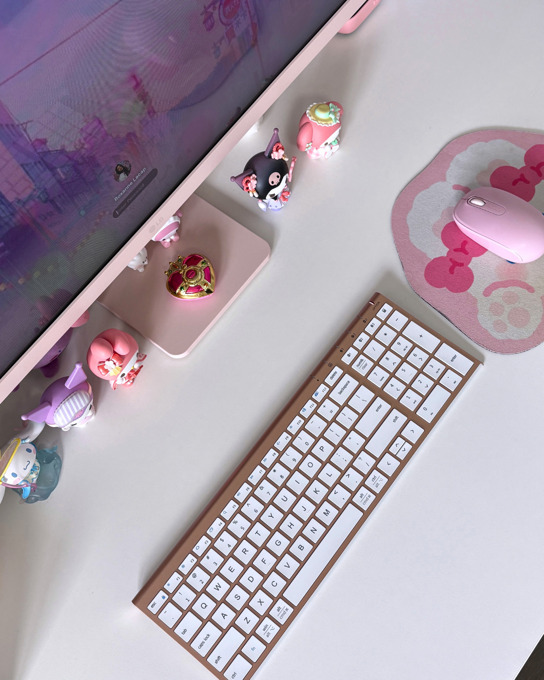 An overhead view of a clean white desk setup featuring a white keyboard with gold trim, a pink mouse on a bow pattern mousepad, and cute Sanrio character figurines arranged neatly around the base of a pink LG monitor.