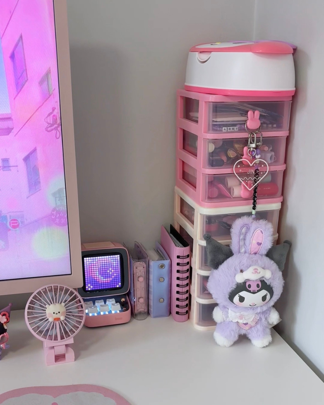 A close-up corner of a kawaii aesthetic desk setup featuring a pink desktop organizer tower, a Divoom pixel art speaker, and a purple Kuromi plush toy next to a pink monitor.