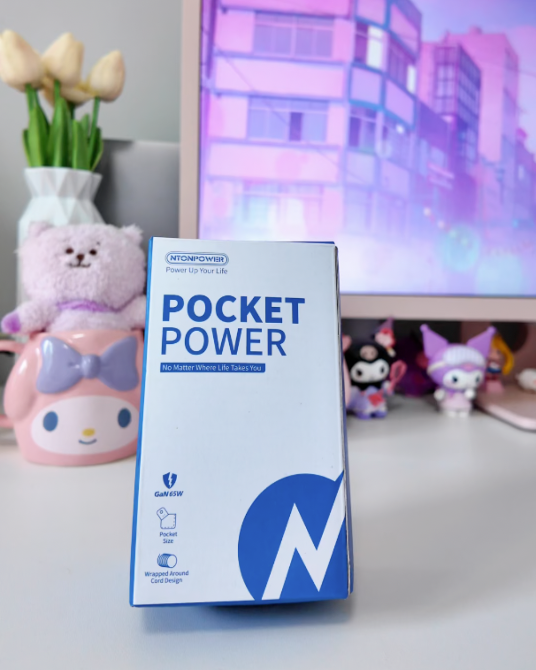 NTONPOWER Pocket Power GaN 65W power strip packaging standing on a white surface as part of a pastel pink and white aesthetic desk setup with a pink monitor and Sanrio plushies.