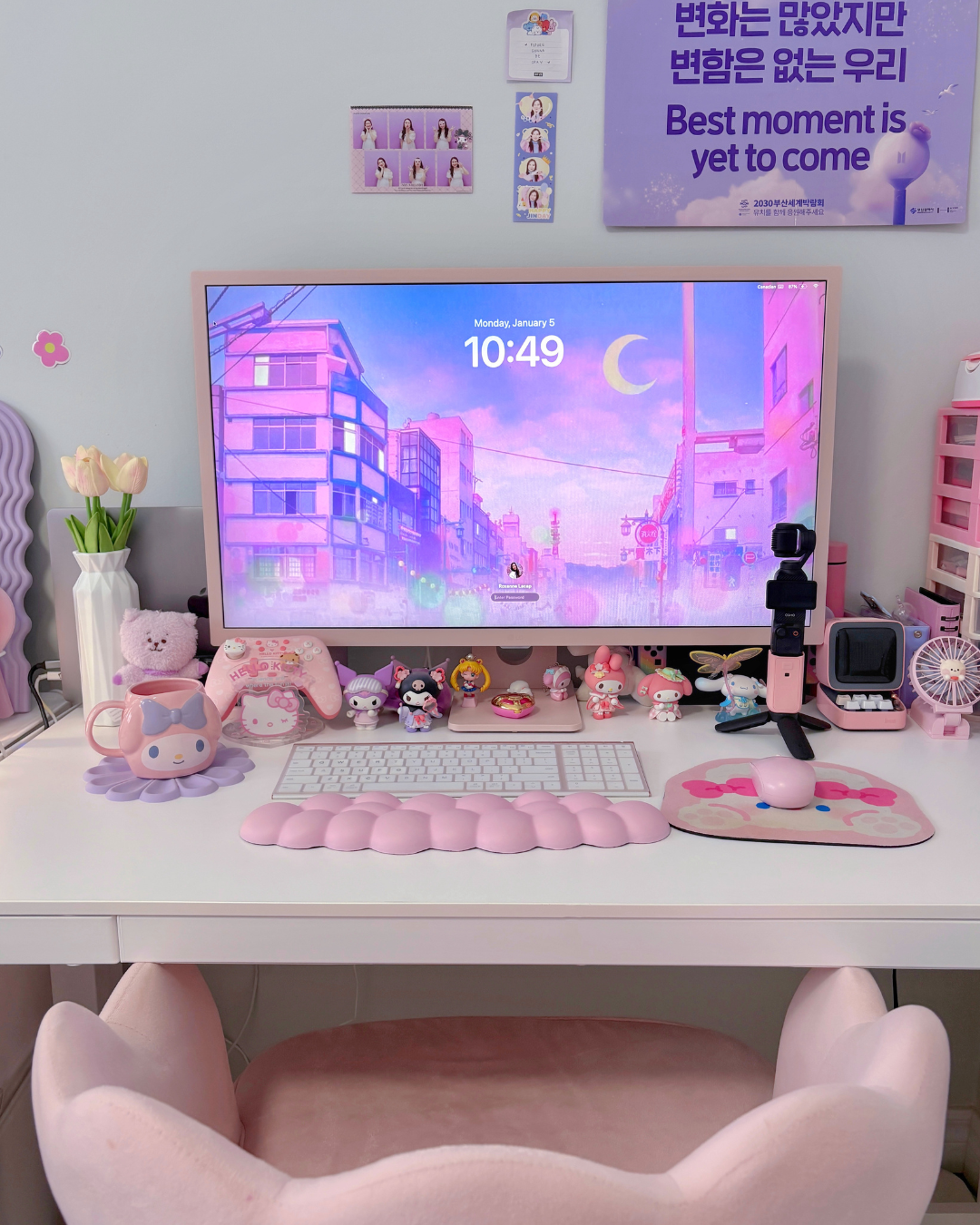 A cozy pink and white aesthetic desk setup featuring a pink desktop monitor with an anime city background, a BTS poster on the wall, a cloud wrist rest, a white keyboard, a pink mouse on a cute mat, a pink desk chair, and various Sanrio character figures lined up under the screen.