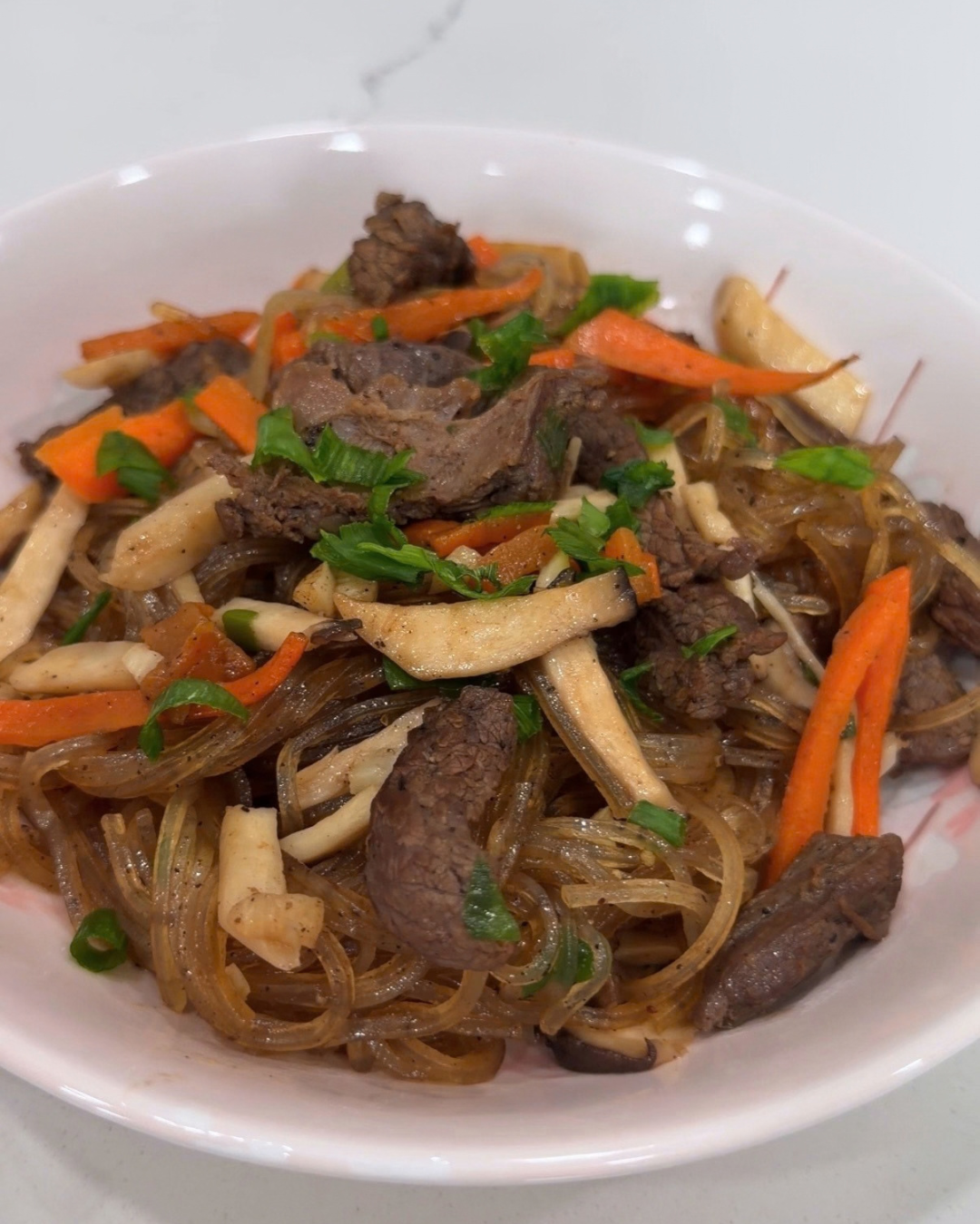 Close-up of homemade Korean Japchae stir-fried noodles topped with tender beef rump, julienned carrots, mushrooms, and green onions.