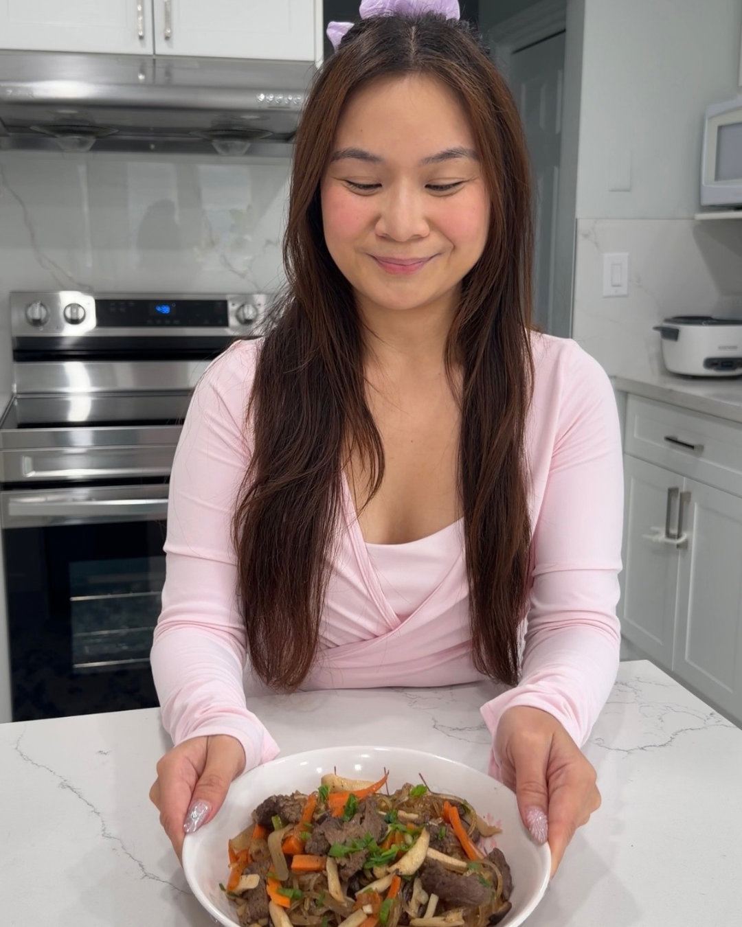 Roxanne presenting a completed bowl of homemade BTS RM-style Japchae on a white kitchen island.